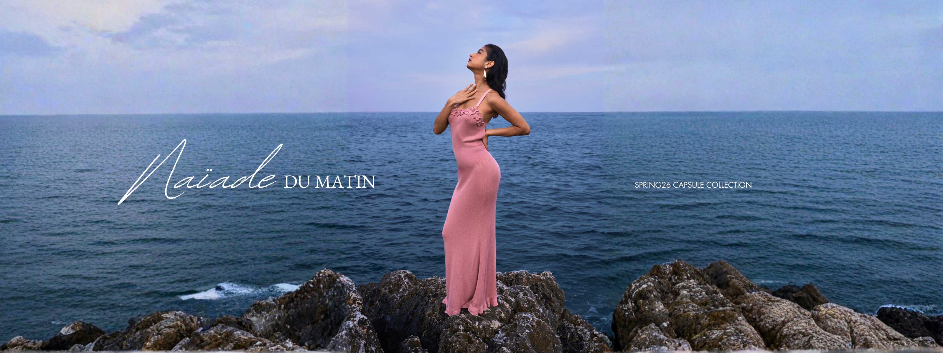 Modèle portant une robe en maille rose ornée de fleurs crochetées à la main par Phème Paris, sur des rochers en bord de mer dans le Sud de la France, évoquant une féminité fluide issue de la collection capsule Printemps Été 2026 ‘Naïade du Matin’.
